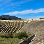 Grande barragem de concreto vista de frente, com reservatório de água e céu azul ao fundo