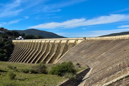 Grande barragem de concreto vista de frente, com reservatório de água e céu azul ao fundo