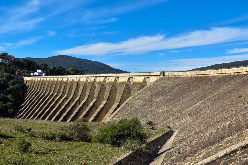 Grande barragem de concreto vista de frente, com reservatório de água e céu azul ao fundo