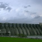 Grande barragem de concreto diante de um céu carregado, destacando sua estrutura e o reservatório de água.