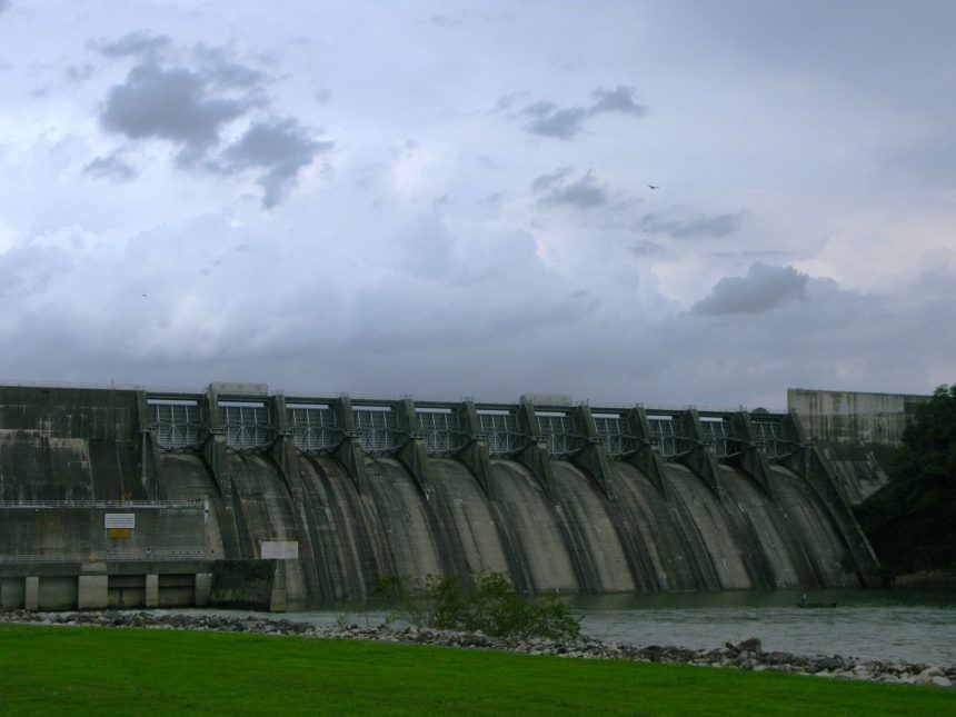 Grande barragem de concreto diante de um céu carregado, destacando sua estrutura e o reservatório de água.